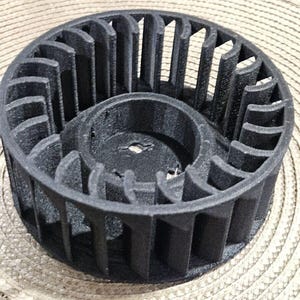 May include: A black, 3D-printed fan impeller with a circular design. The impeller features curved blades and a central hub. The object is made of a matte black material and sits on a beige woven surface.