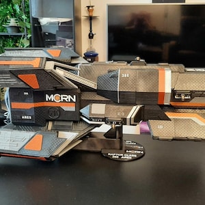 Peut inclure: Maquette détaillée d'un vaisseau spatial gris, noir et orange. Le modèle présente des panneaux complexes et le texte "MCRN ARES NAVY" est visible. Le modèle est exposé sur une surface noire.