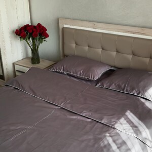 May include: A bed with a grey satin-effect duvet cover and pillowcases with white edging. A vase of red roses sits on a bedside table. The headboard is upholstered in a neutral-toned fabric.