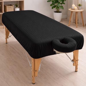May include: A black massage table with a fitted sheet. The sheet has a subtle striped pattern. The table has a face cradle and is supported by wooden legs. The background includes a plant, candles, and a shelf with towels.