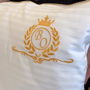 May include: White pillow with a striped pattern and gold embroidered details. The pillow features a gold crown, laurel wreath, and the letters "B" and "O" in a circular design. The edges are trimmed with gold.