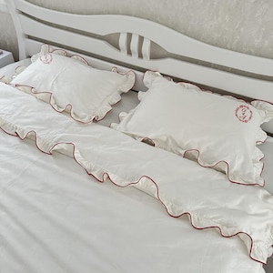 Könnte beinhalten: Weißes Bettwäsche-Set mit roter Bordüre und gestickten Initialen auf den Kissenbezügen. Das Set enthält ein Spannbetttuch, ein Bettlaken und zwei Kissenbezüge. Die Rüschenkanten verleihen der Bettwäsche eine dekorative Note.
