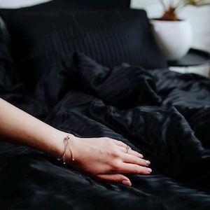 Puede incluir: Primer plano de una cama con ropa de cama negra, que incluye una almohada y sábanas. Una pulsera plateada con esferas colgantes y un anillo adornan una mano que descansa sobre las sábanas. La ropa de cama tiene un sutil estampado de rayas.