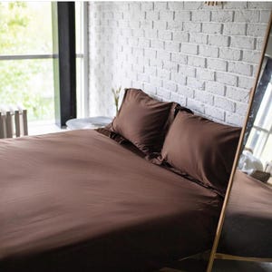 Puede incluir: Un juego de sábanas de color marrón oscuro sólido que incluye dos almohadas rectangulares a juego colocadas cuidadosamente en la cabecera de la cama. La ropa de cama tiene un acabado liso y mate que cubre toda la superficie del colchón.