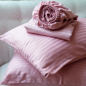 Pode incluir: Um conjunto de roupa de cama rosa, incluindo duas fronhas listradas, um lençol de cima e um lençol com elástico. O lençol com elástico está enrolado. A roupa de cama é de cor rosa lisa com um padrão de listras subtil.