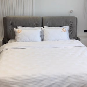 Puede incluir: Una cama con cabecero tapizado gris y ropa de cama blanca. Dos almohadas blancas con un emblema dorado están sobre la cama. El edredón blanco tiene un sutil estampado de rayas. La habitación tiene paredes blancas y suelo de madera clara.