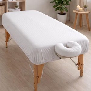 May include: A white massage table with a fitted sheet and a face cradle. The table has wooden legs and is set in a spa-like environment. The sheet has a subtle striped pattern.