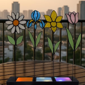 May include: A set of four colourful stained-glass flower garden stakes. The flowers include a daisy, iris, daffodil, and tulip. The stakes are mounted on a black base with three illuminated squares. The background shows a cityscape.