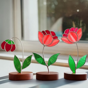 May include: Three stained glass flower sculptures. The flowers have red and green petals and leaves, with silver stems. Each flower is mounted on a small, rectangular wooden base. The background is a window with natural light.