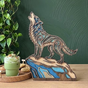 May include: A multi-layered wooden sculpture of a howling wolf standing on a rock formation. The wolf features intricate cut-out patterns with blue and natural wood tones. The rock base is layered with shades of blue and tan, creating a dimensional effect.