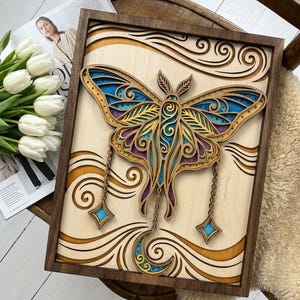 May include: A rectangular wooden wall art piece featuring a layered, laser-cut moth design. The moth has intricate blue, purple, and yellow patterns with dangling star and crescent moon charms. The artwork is set within a dark brown wooden frame.