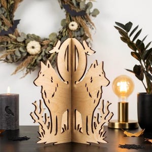 May include: A light-colored wooden decorative piece featuring two symmetrical wolf silhouettes standing amidst stylized tree branches, positioned in front of a circular cutout, creating a three-dimensional structure on a flat surface.