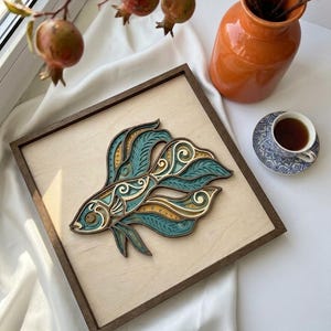 Könnte beinhalten: Ein quadratisches Wandbild aus Holz mit einem mehrschichtigen, lasergeschnittenen Design eines Kampffisches. Der Fisch besteht aus gestapelten Holzschichten in den Farben Petrol, Creme und Naturholz mit komplizierten Wirbelmustern.