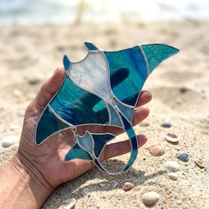 May include: Stained glass manta ray in shades of teal, turquoise, and white, with silver solder lines. The artwork is held against a sandy beach background, showcasing the ocean-inspired design.
