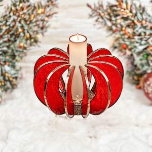 Peut inclure: Un bougeoir rouge et argenté avec une bougie blanche allumée. Le bougeoir a un design circulaire avec des panneaux rouges et des bordures argentées. L'arrière-plan est flou avec des branches de sapin de Noël.