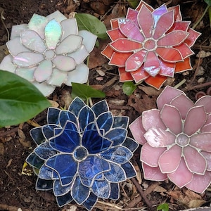 May include: Four colorful stained glass flower ornaments in white, red-orange, blue, and pink. Each flower has multiple petals and a silver-colored border. The flowers are arranged on a bed of soil and leaves.