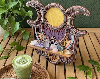 May include: A layered wooden wall shelf featuring a triple moon design with a central yellow sun disc, purple and dark wood accents, and a small horizontal ledge holding an assortment of raw and polished crystals including quartz and amethyst.
