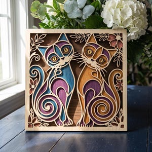 May include: A square layered wooden wall art piece featuring two stylized cats with intricate cutouts. The left cat is blue and purple, while the right cat is orange and purple. The design includes floral accents and swirling patterns, all set within a light-colored wooden frame.