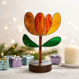 May include: A stained glass flower sculpture with orange, yellow, and red petals, green leaves, and a brown stem. The flower is mounted on a round, dark brown wooden base. The background includes a lit candle and small wrapped gifts.