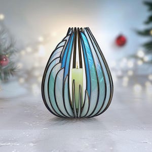 May include: A decorative candle holder with a blue and white stained glass design. The holder has a teardrop shape with a black metal frame and a lit green candle inside. The background is blurred with Christmas decorations.