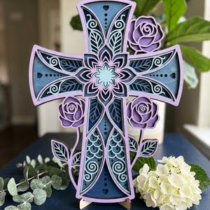 May include: A multi-layered wooden cross featuring intricate laser-cut designs in shades of purple, lavender, and blue. The cross is decorated with floral rose motifs and mandala-inspired patterns, standing upright on a small wooden base.