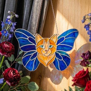 Puede incluir: Gato de vidriera con alas de mariposa, con cuerpo naranja y amarillo y alas azules. El gato tiene ojos turquesa y está suspendido por una cadena plateada. El fondo incluye flores y una cortina oscura.
