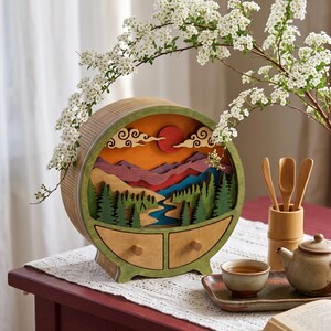May include: A circular wooden storage box with two small drawers, featuring a layered landscape scene of green pine trees, a blue river, purple mountains, and a red sun under stylized clouds, framed by a green border and light wood grain.