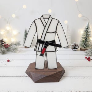 Może przedstawiać: Szkło artystyczne przedstawiające biały strój sztuk walki z czarnym pasem i czerwonym paskiem. Strój jest zamontowany na ciemnobrązowej, geometrycznej drewnianej podstawie. W tle dekoracje świąteczne.