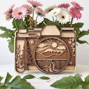 May include: A multi-layered wooden wall art piece shaped like a camera. The design features a landscape scene within the lens area, including a sun, clouds, mountains, and a river. A tall pine tree is on the left, with smaller trees on top.