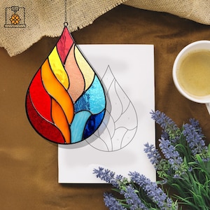 May include: A vibrant stained glass teardrop suncatcher in shades of red, orange, yellow, and blue. The suncatcher is suspended by a chain. A drawing of the same shape, a cup of coffee, and lavender sprigs are also visible in the image.