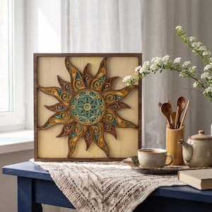 May include: A square wooden wall art piece featuring a multi-layered sun design with intricate swirling patterns in shades of teal, orange, and natural wood tones, framed within a dark brown border and set against a light wood background.