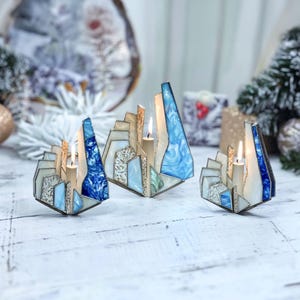 May include: Three decorative candle holders with a stained glass design. The holders feature a geometric shape with shades of blue, white, and beige. Each holder has a lit candle inside, creating a warm glow.
