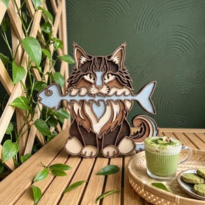 May include: A multi-layered wooden wall art piece depicting a cat with tufted ears holding a light blue fish skeleton in its mouth. The cat features various shades of brown, cream, and light blue wood, creating a dimensional, stacked appearance.