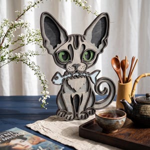 May include: A multi-layered wooden cat sculpture featuring shades of grey, large green eyes, and a light blue fish held in its mouth. The cat has pointed ears, a curled tail, and a detailed cutout design that creates depth through stacked wood layers.