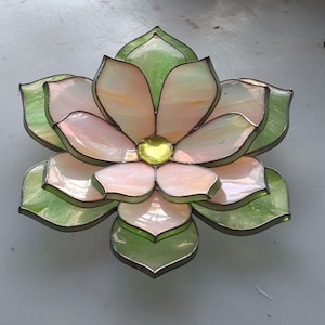 May include: A stained glass flower in shades of pink, green, and yellow. The petals are a soft pink with green leaves and a yellow centre. This decorative piece is likely for home decor.