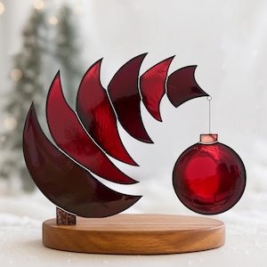 Op de afbeelding: Een decoratieve kerstversiering met een reeks rode, halvemaanvormige elementen in een spiraal, en een hangende rode bal. De versiering is gemonteerd op een ronde houten basis, wat een feestelijke uitstraling creëert. De basis is ongeveer 10 cm in diameter.