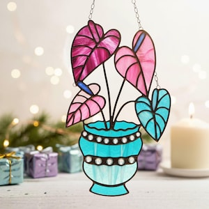 Puede incluir: Arte de vidriera de una planta con hojas rosas y azules en una maceta turquesa. Las hojas tienen forma de corazón y están conectadas por tallos negros. La maceta tiene un borde decorativo con detalles blancos. La obra cuelga de una cadena.
