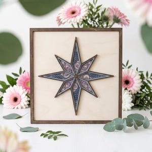 May include: A square wooden wall art piece featuring a layered, multi-dimensional eight-pointed star in shades of purple, blue, and light wood. The star is centered on a light-colored wooden panel framed by a dark brown wooden border.