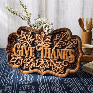 May include: A multi-layered wooden sign featuring the text GIVE THANKS in a central cutout, surrounded by intricate laser-cut maple leaf patterns and decorative scrollwork in shades of natural wood, dark brown, and deep blue.