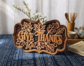 Può includere: Un'insegna in legno a più strati con il testo GIVE THANKS in un intaglio centrale, circondato da intricati motivi di foglie d'acero tagliate al laser e volute decorative in tonalità di legno naturale, marrone scuro e blu intenso.