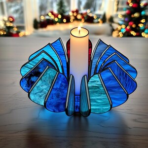 Peut inclure: Un bougeoir en vitrail avec une bougie blanche allumée. Le bougeoir a un design en forme d'éventail avec des nuances de verre bleu et turquoise. Le bougeoir est posé sur une surface en bois, avec des décorations de Noël en arrière-plan.