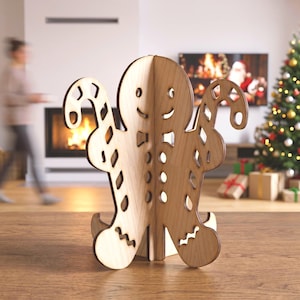 May include: A light brown wooden gingerbread person decoration featuring a cutout design. The figure has a smiling face, a bow tie, and holds a candy cane in each hand. The structure is composed of interlocking flat wooden pieces that allow it to stand upright on a flat surface.