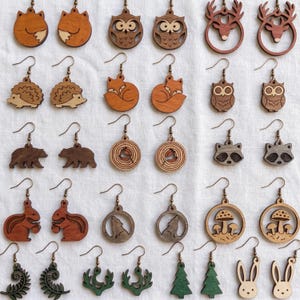 May include: A collection of thirty pairs of wooden earrings featuring various nature-inspired designs including foxes, owls, deer, hedgehogs, bears, raccoons, squirrels, wolves, mushrooms, ferns, pine trees, and rabbits, all with metal earring hooks.