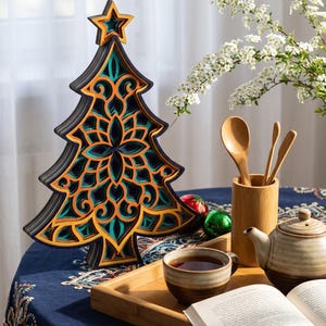 May include: A multi-layered wooden Christmas tree decoration featuring intricate cut-out patterns in shades of dark blue, bright orange, and teal. The tree is topped with a star and displays a complex geometric design throughout its structure.