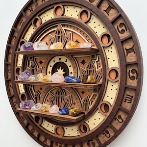 May include: A circular wooden wall shelf featuring three horizontal tiers for display. The frame is carved with twelve zodiac symbols. The background displays layered wood cutouts of crystals and stars. Various polished gemstones sit on the shelves.