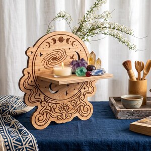 May include: A light brown wooden wall shelf featuring laser-cut engravings of a crystal ball, moon phases, stars, and an eye. The shelf holds a small lit candle and a collection of various colorful raw crystals and polished gemstones.