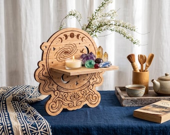 May include: A light brown wooden wall shelf featuring laser-cut engravings of a crystal ball, moon phases, stars, and an eye. The shelf holds a small lit candle and a collection of various colorful raw crystals and polished gemstones.