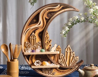 May include: A crescent moon-shaped wooden wall shelf featuring layered cutouts, crystal-shaped accents, and a small cat silhouette. The shelf includes two horizontal tiers holding various small polished stones and crystals.