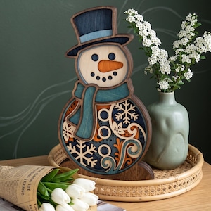 May include: A layered wooden snowman decoration featuring a dark blue top hat, a teal scarf, and an orange carrot nose. The body displays intricate cutouts of snowflakes and swirling patterns in shades of blue, orange, and natural wood tones.