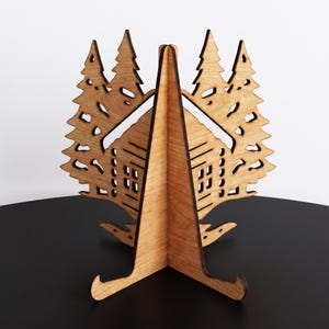 May include: A light brown wooden display stand featuring a laser-cut design of a cabin nestled among pine trees. The structure consists of two interlocking pieces that form a cross-shaped base with curved supports at the bottom for holding items.